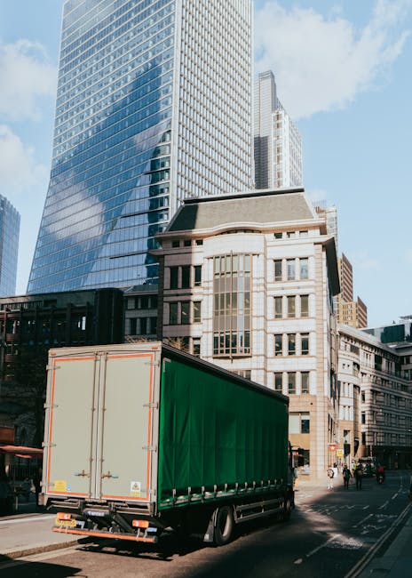 A burgundy light commercial pickup truck parked on a city street in front of a modern office building with large glass windows and white concrete panels. The truck's flatbed is loaded with various pac